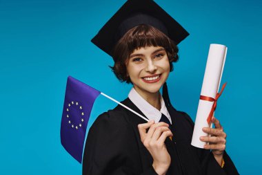 Mavi zemin üzerinde diploma ve AB bayrağı bulunan önlüklü ve akademik şapkalı mutlu üniversite mezunu kız