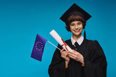 Mavi zemin üzerinde diploma ve AB bayrağı bulunan, önlüklü ve akademik şapkalı üniversiteli neşeli mezun kız