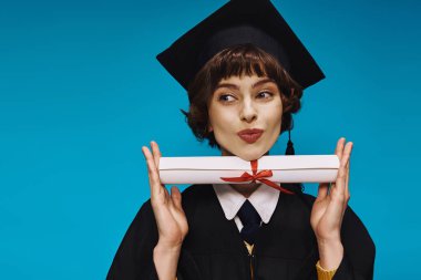 Gecelikli, akademik şapkalı, dudak büken ve diplomasını gururla tutan mutlu üniversiteli kız.