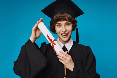 Gecelikli ve akademik şapkalı neşeli üniversiteli kız mavi diplomasını gururla tutuyor, mezun oluyor.