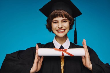 Mavi elbisesi ve akademik şapkası olan liseli kız diplomasını gururla tutuyor.