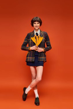 full length of stylish student in checkered blazer and skirt on orange background, college uniform clipart