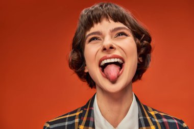 Playful female student in college uniform sticking out tongue, lively on orange background clipart
