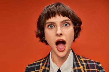 Surprised college girl looking at camera with wide eyes on orange backdrop, face expression clipart