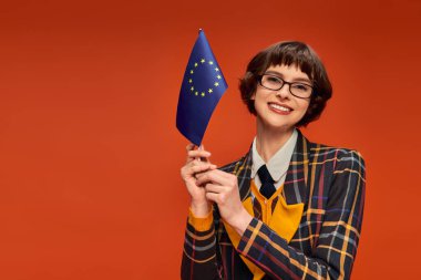 happy young college girl in uniform and glasses holding EU flag on vibrant orange background clipart