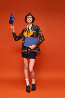 happy young college girl in uniform and glasses holding EU flag with notebook on orange background clipart