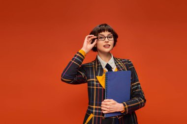 Kolej üniformalı düşünceli bir öğrenci gözlüğünü ayarlıyor ve elinde turuncu arka planda defter tutuyor.