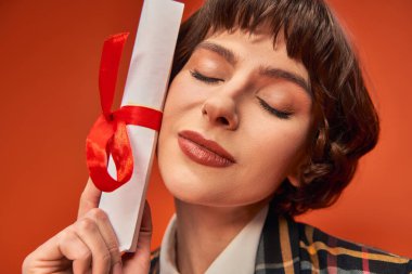 portrait of smiling college girl in uniform holding her graduation diploma on orange backdrop clipart