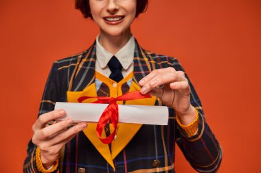 Kırpılmış mutlu üniversiteli üniformalı kız elinde turuncu arka planda mezuniyet diploması tutuyor.