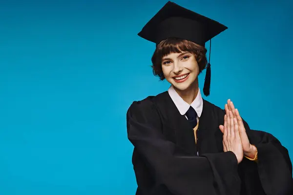 cheerful young woman in black graduation gown and academic cap smiling on blue background, ceremony