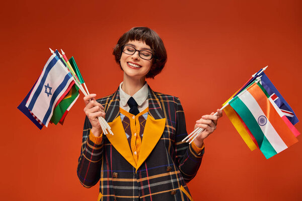 happy college girl in her uniform and glasses holding multiple flags and standing on orange backdrop
