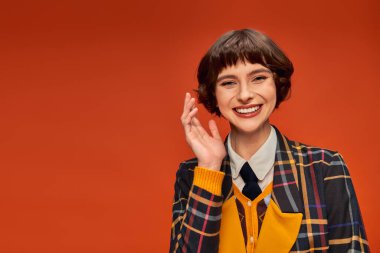 optimistic college girl in checkered uniform waving hand on orange background, happy student life clipart