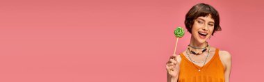 Turuncu örgü bluzlu neşeli genç kadın elinde pembe pankartta renkli tatlı lolipoplar tutuyor.