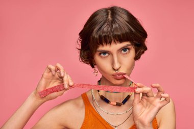 Esmer ve burnu delen genç bir kadın pembe arka planda tatlı ve ekşi şeker şeridi ısırıyor.