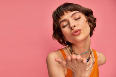 flirty young woman with nose piercing and short brunette hair showing kiss on pink background clipart