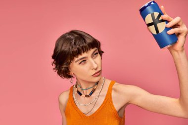 doubtful young woman with short brunette hair checking out her soda can on pink, summer drink clipart