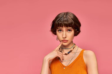 20 'li yaşlardaki endişeli genç bir kadın pembe arka planda turuncu örülü kolsuz bir bluzla dikiliyor.