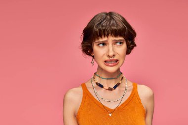 puzzled young girl in her 20s standing in orange knitted tank top on pink background, unsure clipart