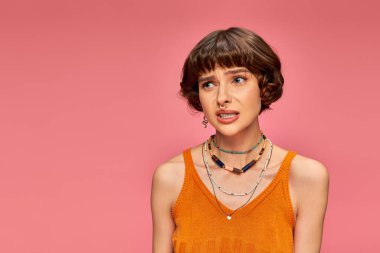 puzzled young woman in her 20s standing in orange knitted tank top on pink background, uneasy clipart