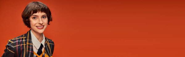 оптимистичная и молодая студентка в клетчатой униформе колледжа улыбается на оранжевом фоне, баннер