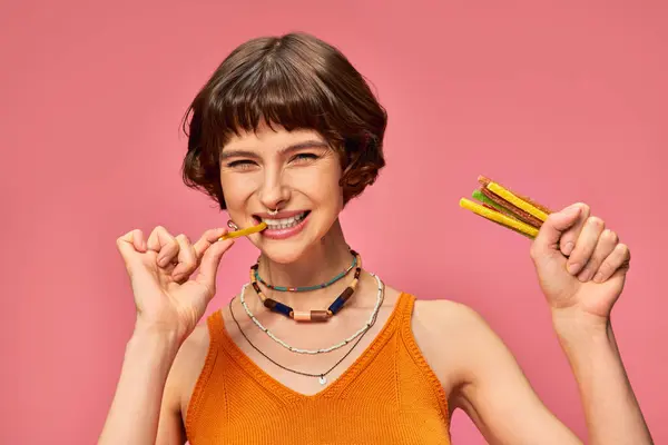 20代の若いピアスされた女性は甘くて酸っぱいガミーのストライプを噛み ピンクでキャンディーを保持 — ストック写真