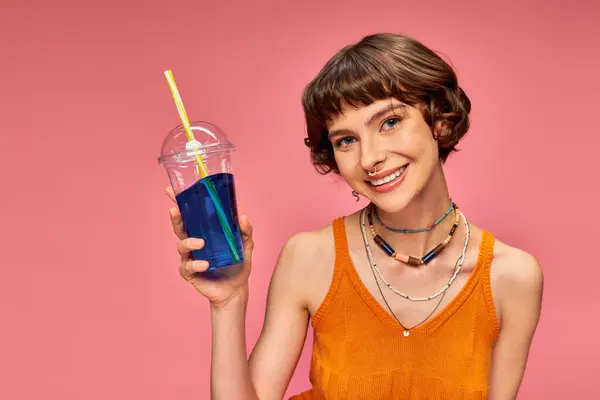 ピンクの背景に爽やかな夏の飲み物を保持する短いブルネット髪の肯定的な若い女性 — ストック写真