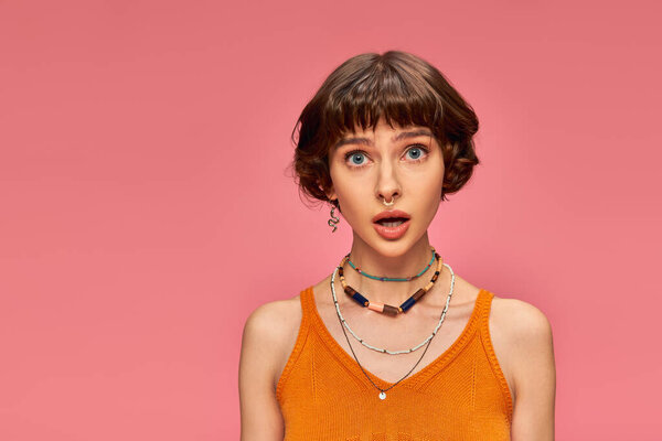 astonished young girl in her 20s standing in orange knitted tank top on pink background, disbelief