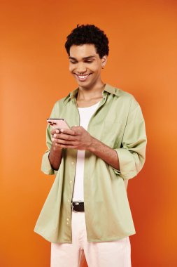 Keyifli Afro-Amerikan erkek rahat giyinmiş turuncu arka plandaki akıllı telefonuna bakıyor.