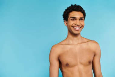 cheerful handsome african american man in blue swimming trunks looking away on blue backdrop clipart