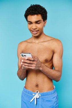 appealing jolly african american man in blue swimming trunks looking at phone on blue backdrop clipart