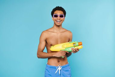 joyful young african american man in swimming trunks with stylish sunglasses posing with water gun clipart