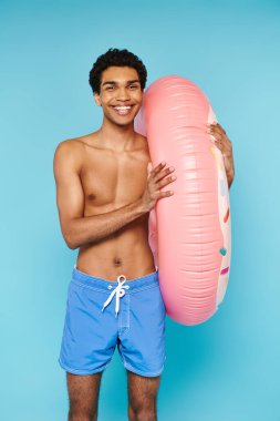 joyous african american man posing with swimming ring on blue backdrop and looking at camera clipart