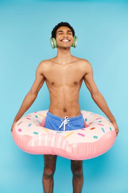 Yüzme şortlu neşeli Afro-Amerikalı adam şişme donutla kulaklıkla müzik dinliyor.