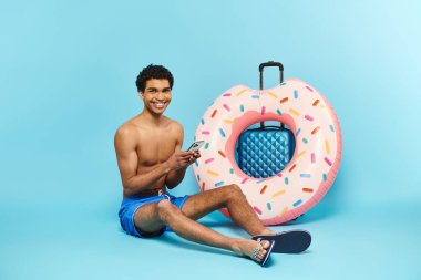 Neşeli Afrikalı Amerikalı adam akıllı telefonunu tutuyor ve bavulun yanında oturuyor ve şişirilebilir donut yiyor.