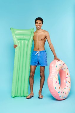 Neşeli Afrikalı Amerikalı adam şişirilebilir donut ve şişme yatakla poz veriyor ve kameraya gülümsüyor.