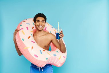 Neşeli Afro-Amerikalı adam şişme donutla mavi arka planda ferahlatıcı kokteyllerin tadını çıkarıyor.