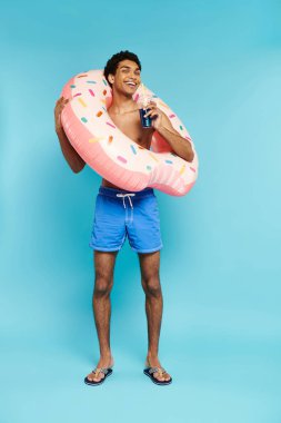 Neşeli Afrikalı Amerikalı adam şişme donutla mavi arka planda ferahlatıcı kokteyllerin tadını çıkarıyor.