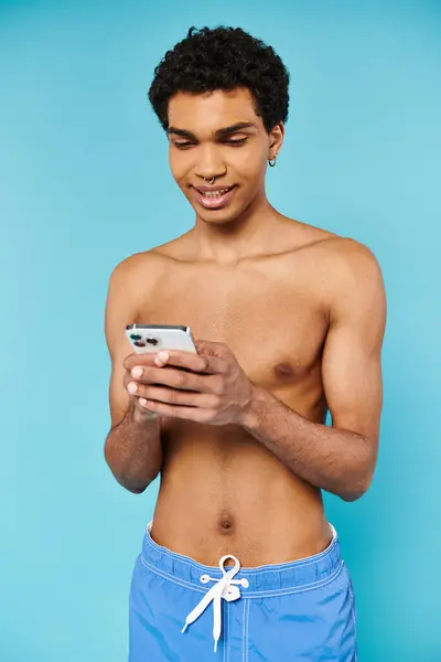 stock image appealing jolly african american man in blue swimming trunks looking at phone on blue backdrop