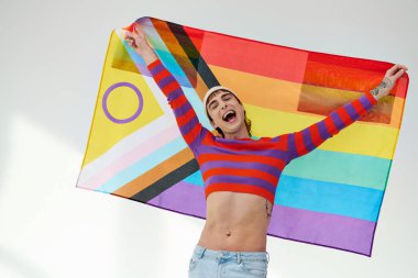 joyous appealing gay man in vivid outfit with white hat holding rainbow flag and looking at camera clipart