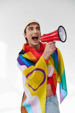 Gökkuşağı bayrağıyla canlı giyinmiş neşeli, şık bir gay adam megafon kullanıyor ve başka tarafa bakıyor.