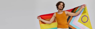 Neşeli, siyah saçlı, elinde gökkuşağı bayrağı olan ve kameraya mutlu bir şekilde gülümseyen neşeli bir gay.