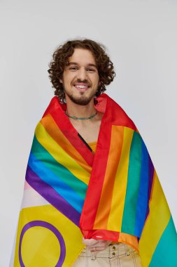 Neşeli, çekici, siyah saçlı, elinde gökkuşağı bayrağı olan ve kameraya mutlu bir şekilde gülümseyen gay adam.