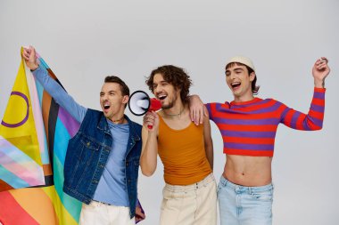 Gökkuşağı bayrağı taşıyan ve gri arka planda megafon kullanan rahat giyimli üç neşeli gay adam.