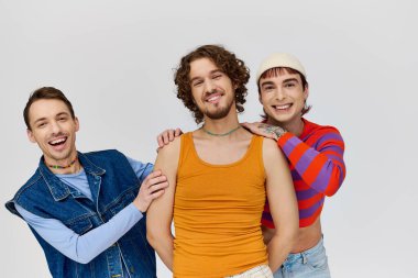 Canlı giyinmiş neşeli genç gay adamlar gri arka planda birbirlerine poz verip kameraya bakıyorlar.
