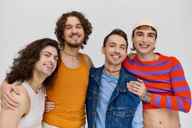 Gri arka planda poz verirken kameraya gülümseyen canlı giyinmiş dört neşeli yakışıklı gay adam.