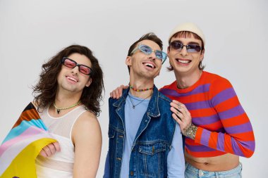 Üç çekici neşeli gay adam şık güneş gözlükleriyle gökkuşağı bayrağıyla poz veriyor ve başka tarafa bakıyor.