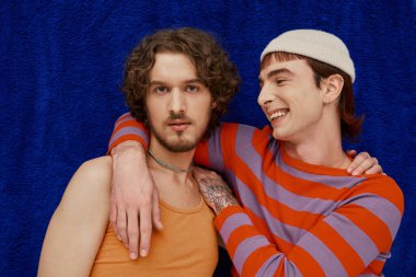 two joyful good looking gay men in vibrant clothes posing on dark blue backdrop, pride month clipart