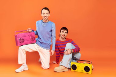 Canlı makyajlı, turuncu arka planda kasetçalarla poz veren neşeli, yakışıklı iki gay adam.