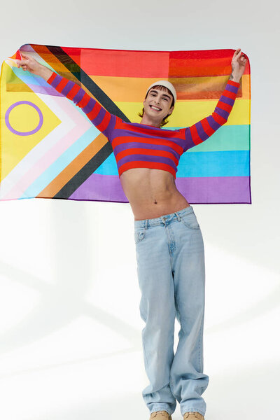 positive appealing gay man in vivid outfit with white hat holding rainbow flag and looking at camera