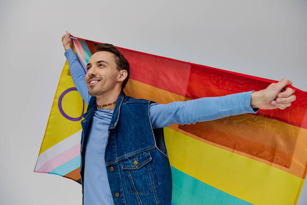 cheerful handsome gay man in vivid trendy outfit holding rainbow flag and looking away, pride month